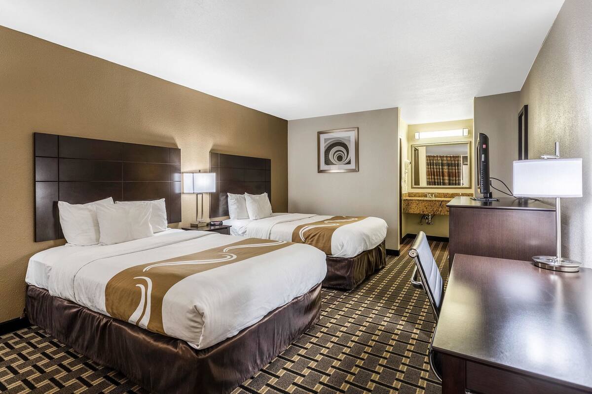 A hotel room with two beds, a desk, a mirror, and a bathroom visible through an open door.