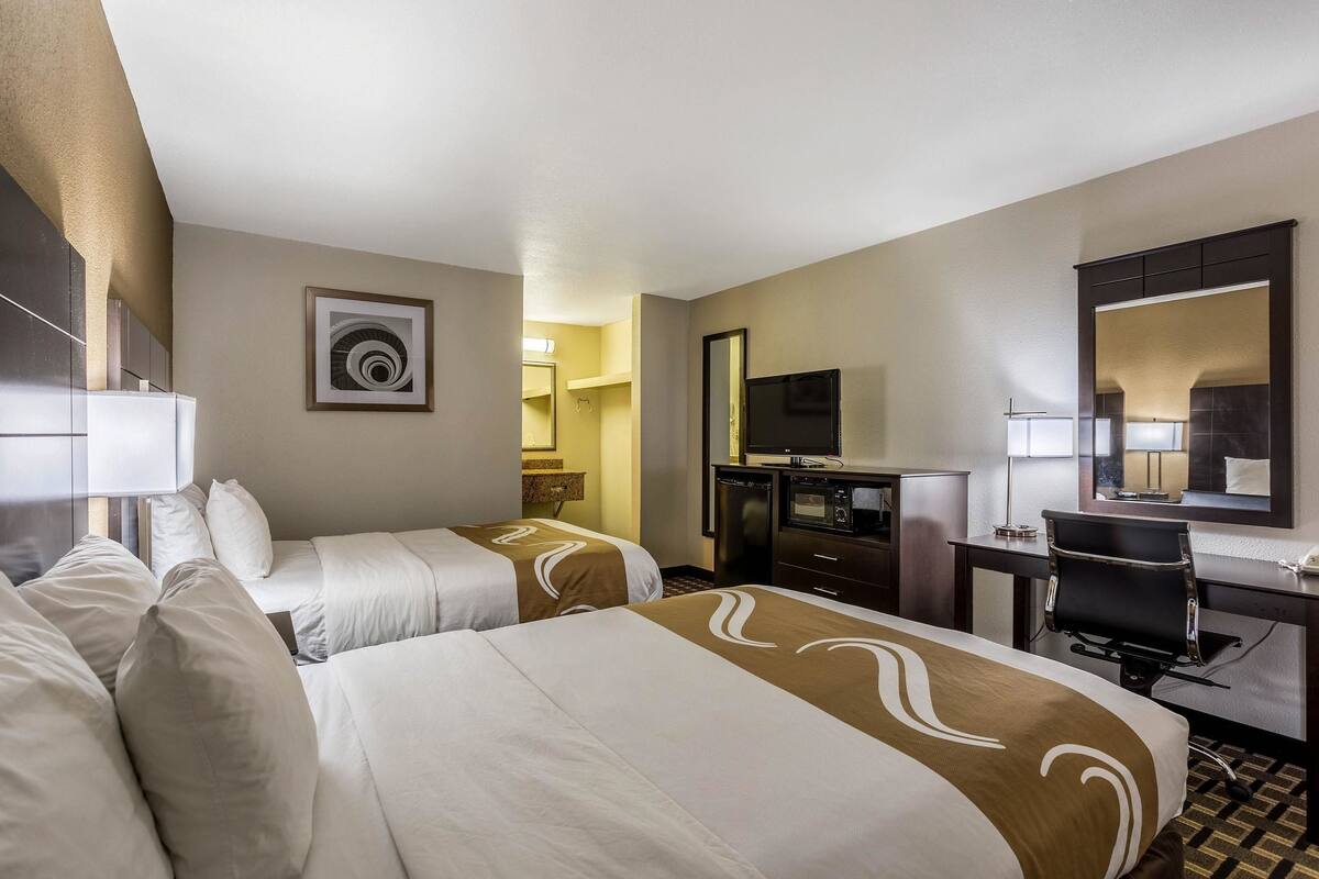 A hotel room with two beds, a desk, a chair, a TV, and a bathroom visible in the background.