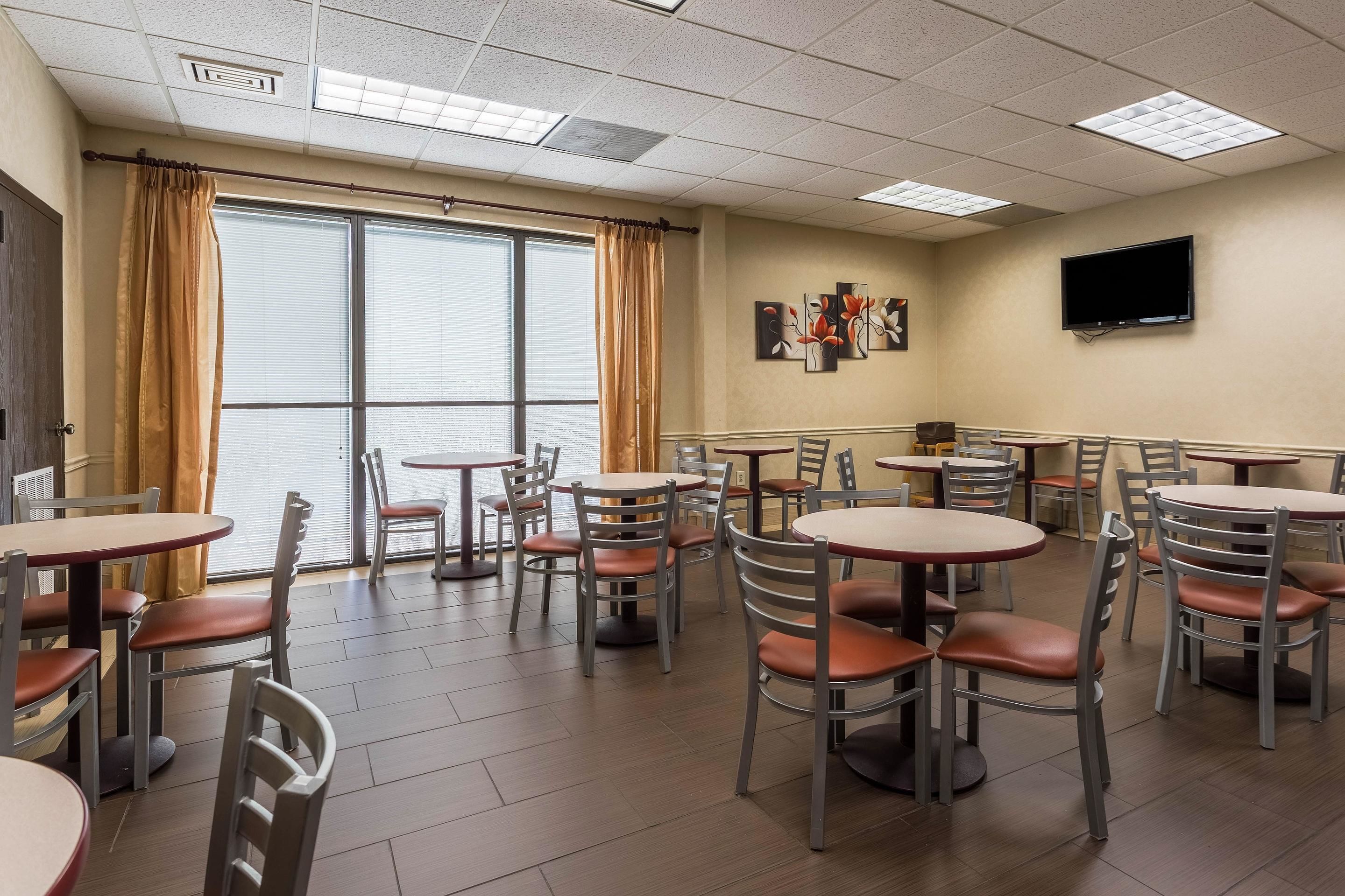 A dining area with multiple round tables and chairs, a television mounted on the wall, and large windows with curtains.