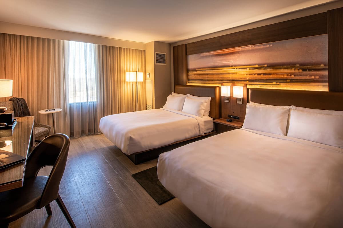A hotel room with two beds, a desk, a chair, and a large window with curtains.