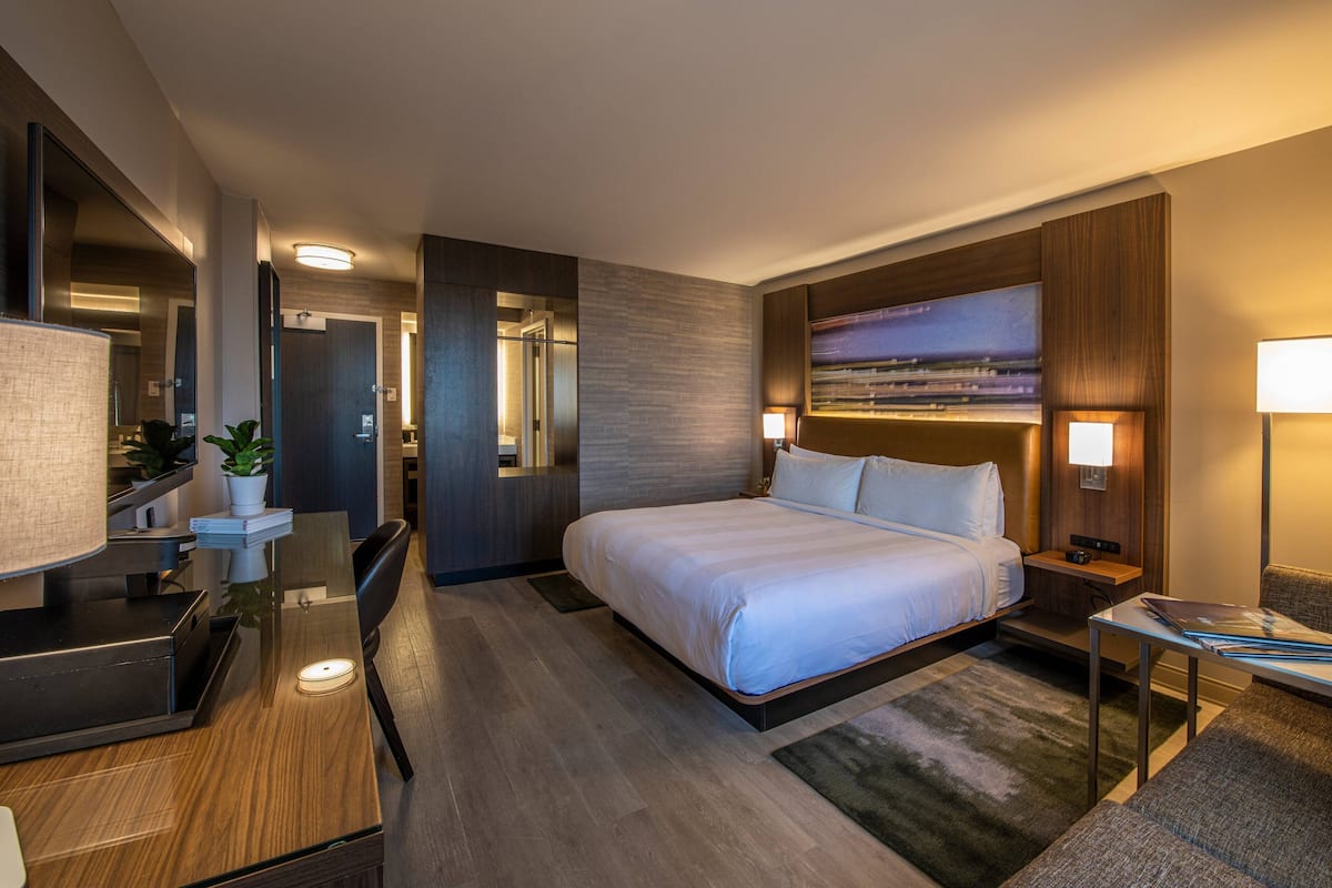 A modern hotel room with a large bed, a desk with a lamp, a mirror, and a painting on the wall.
