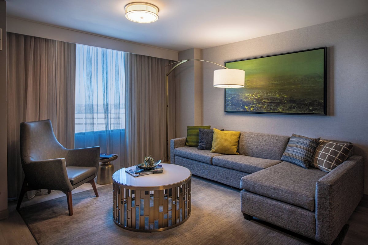 A modern hotel room with a grey sofa, a round coffee table, a leather armchair, and a large window with curtains.