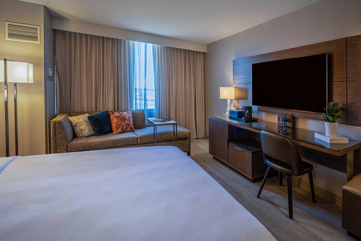 A hotel room with a large bed, a sofa with patterned cushions, a desk with a computer, a flat-screen TV, and a window with curtains.