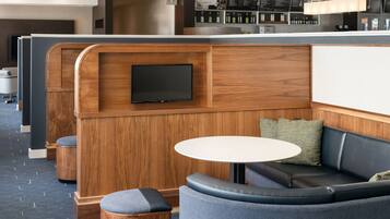 A modern hotel lobby with a wooden reception desk, a flat-screen TV, a round table, and a seating area with a sofa and armchairs.