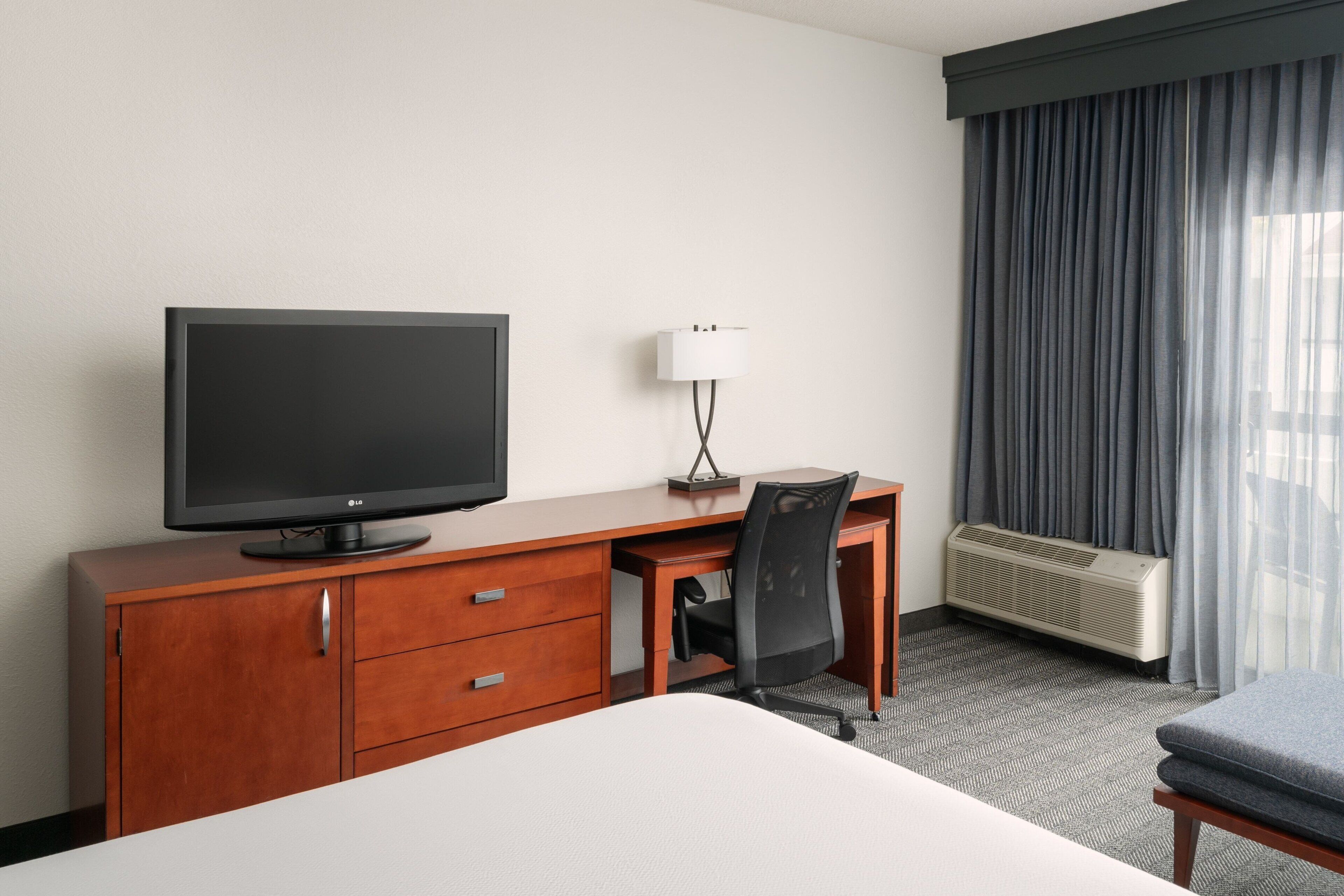 Habitación de hotel con cama, escritorio con televisor, silla, lámpara y ventana con cortinas.