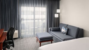 Habitación de hotel con sofá gris, una mesa de centro de madera, cama, escritorio con silla y un ventanal con cortinas.
