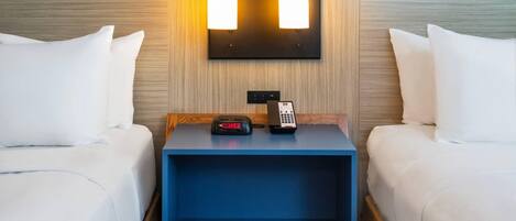 A hotel room with two beds, a nightstand with a clock and phone, and a wall-mounted light fixture.