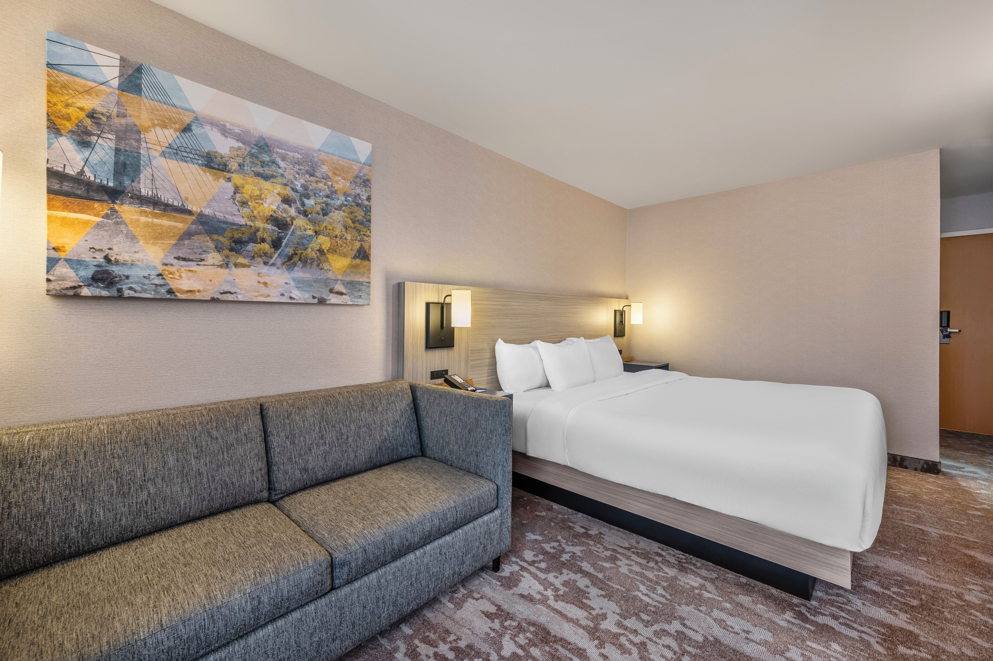 A hotel room with a large bed, a sofa, a wall-mounted artwork, and a nightstand with a lamp.