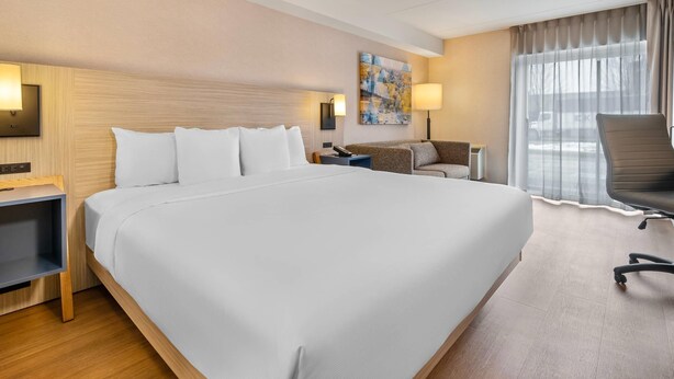 Habitación de hotel con una cama grande, un escritorio, una silla, una lámpara, un cuadro en la pared y una ventana con cortinas.