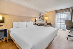 Habitación de hotel con una cama grande, un escritorio, una silla, una lámpara, un cuadro en la pared y una ventana con cortinas.