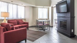 Una sala de estar moderna con un sofá rojo, una mesa de comedor con sillas, un televisor de pantalla plana sobre una chimenea y ventanas grandes con cortinas blancas.