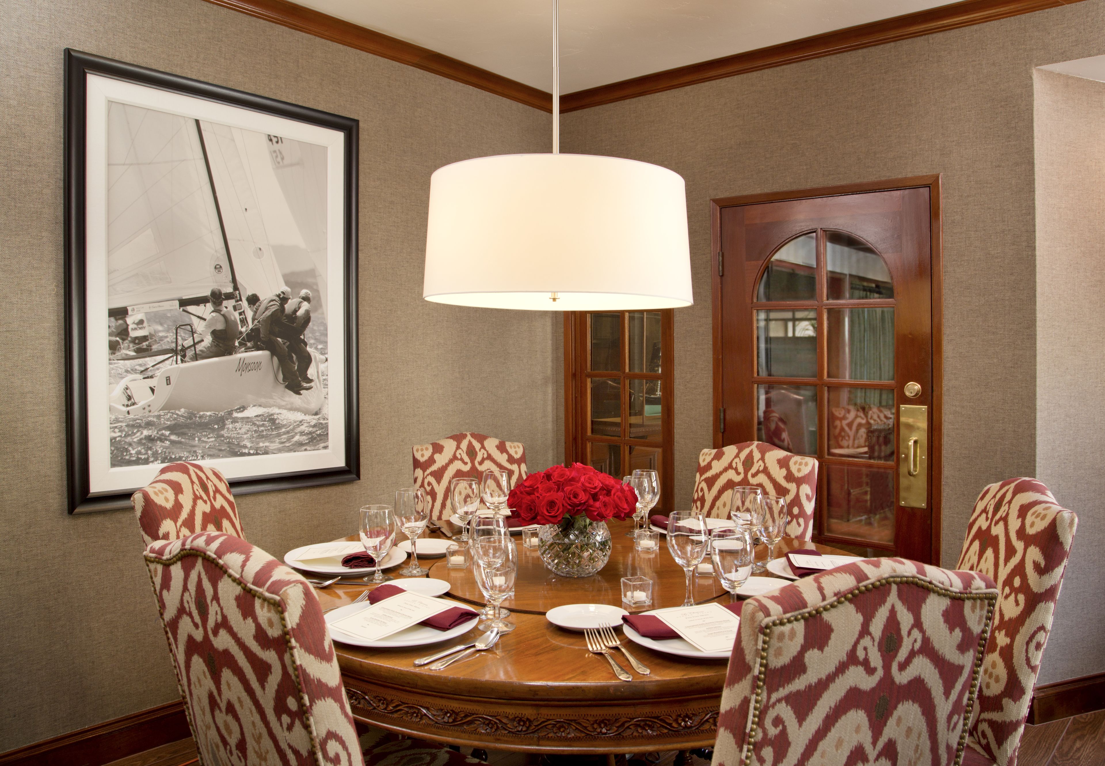 Comedor con una mesa redonda dispuesta para seis personas, sillas estampadas, un jarrón con rosas rojas y una fotografía en blanco y negro enmarcada en la pared.