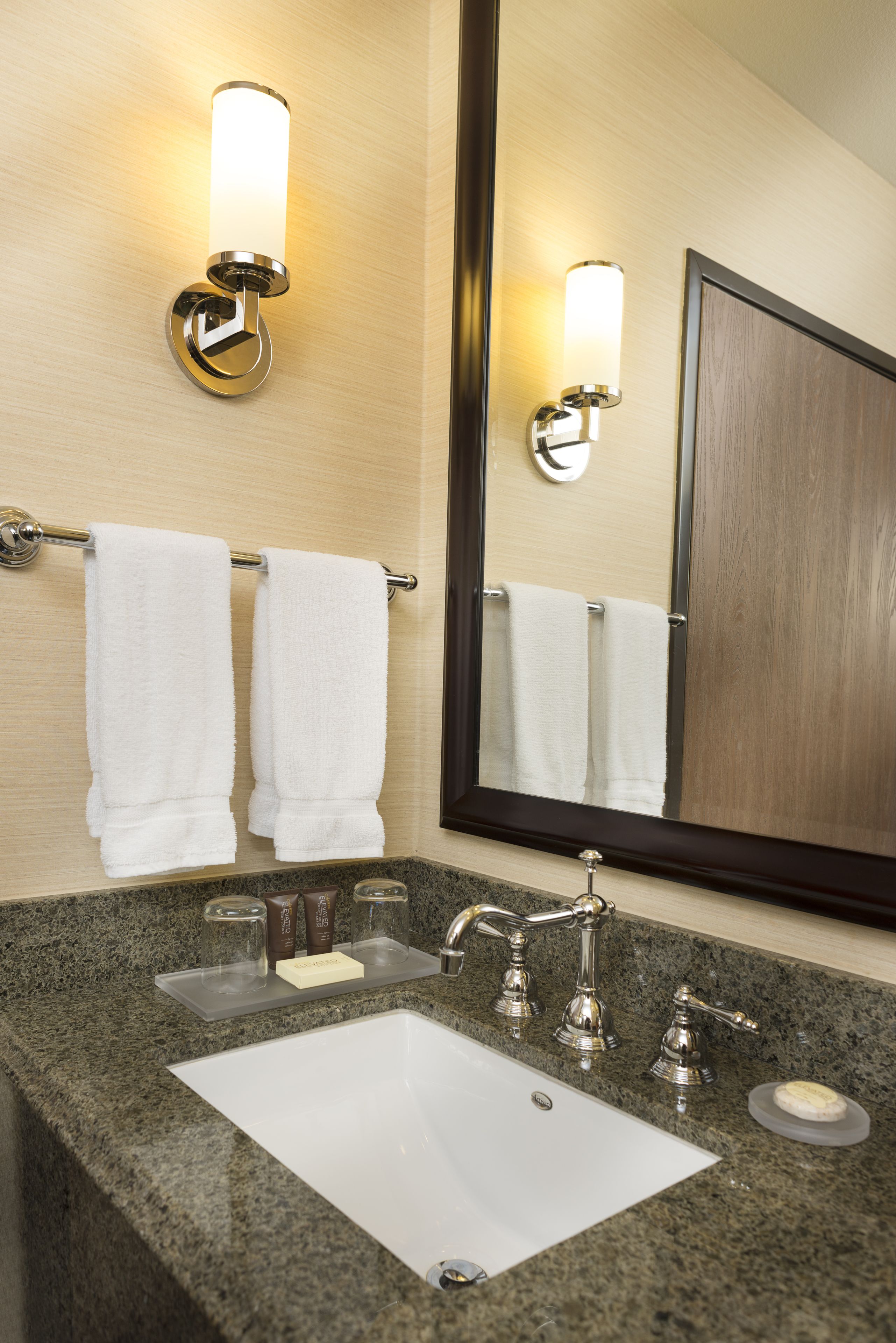 A bathroom with a granite countertop, a white sink, two wall-mounted towel racks, a mirror, and two wall sconces.