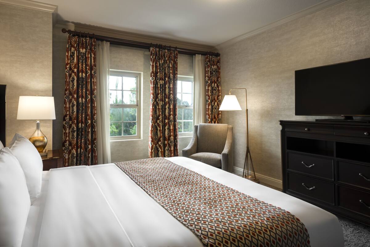 A hotel room with a large bed, two windows with curtains, a chair, a TV, and a lamp.