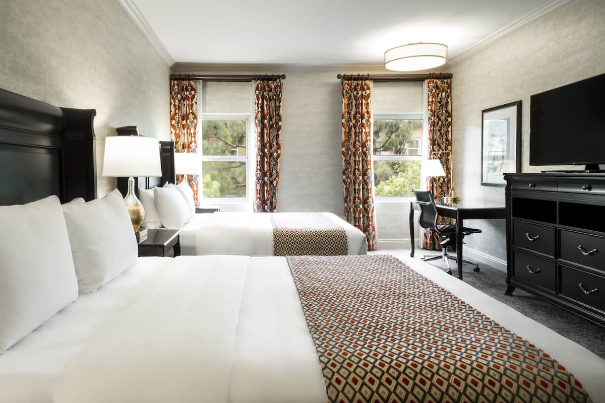 A hotel room with two beds, a desk, a TV, and a large window with curtains.