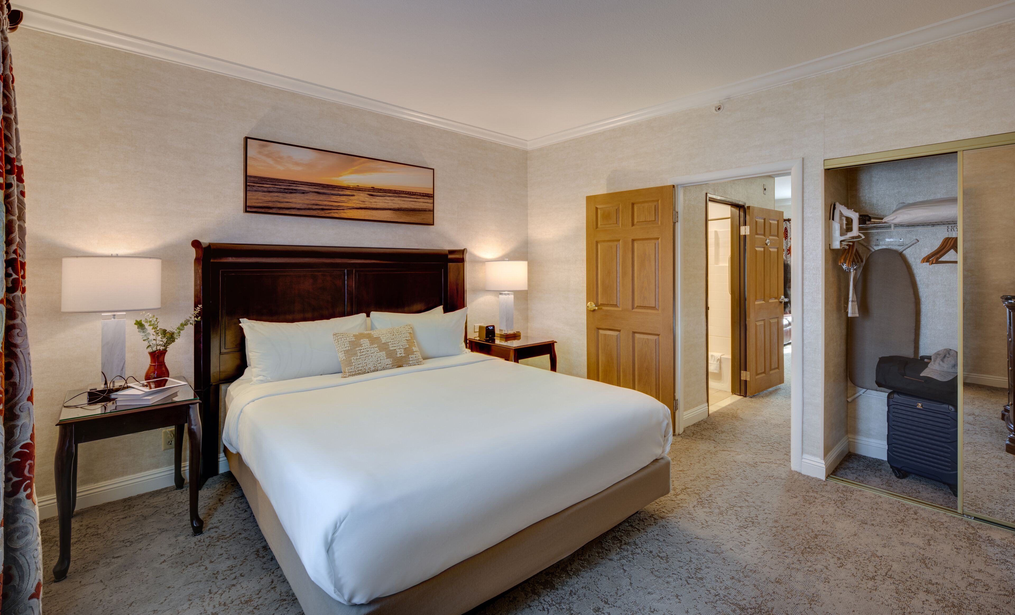 A hotel room with a large bed, two bedside tables, a nightstand, a painting above the bed, and a wardrobe with hanging clothes.