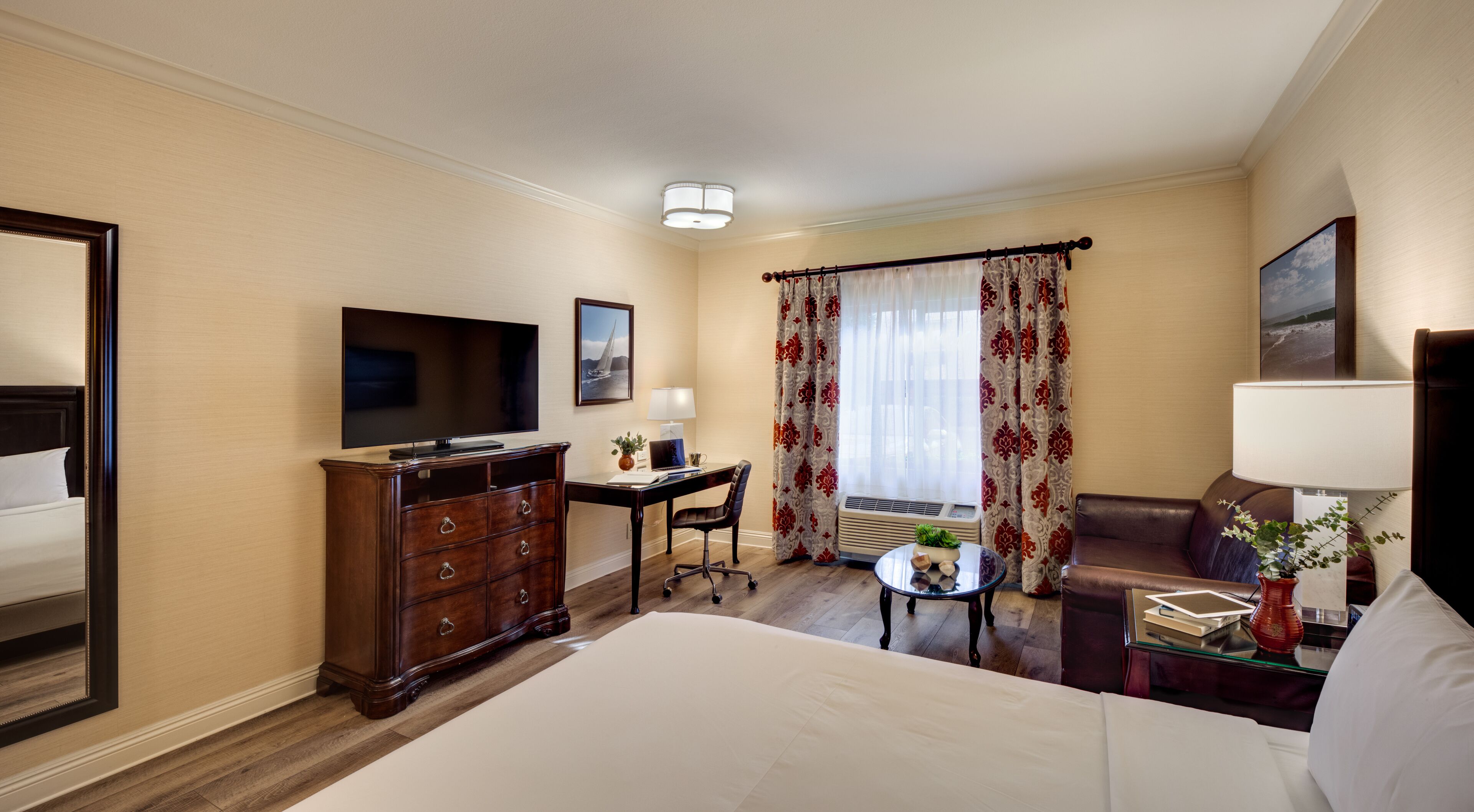 Een hotelkamer met een bed, een bureau, een stoel, een televisie, een lamp, een raam met gordijnen en een plant.