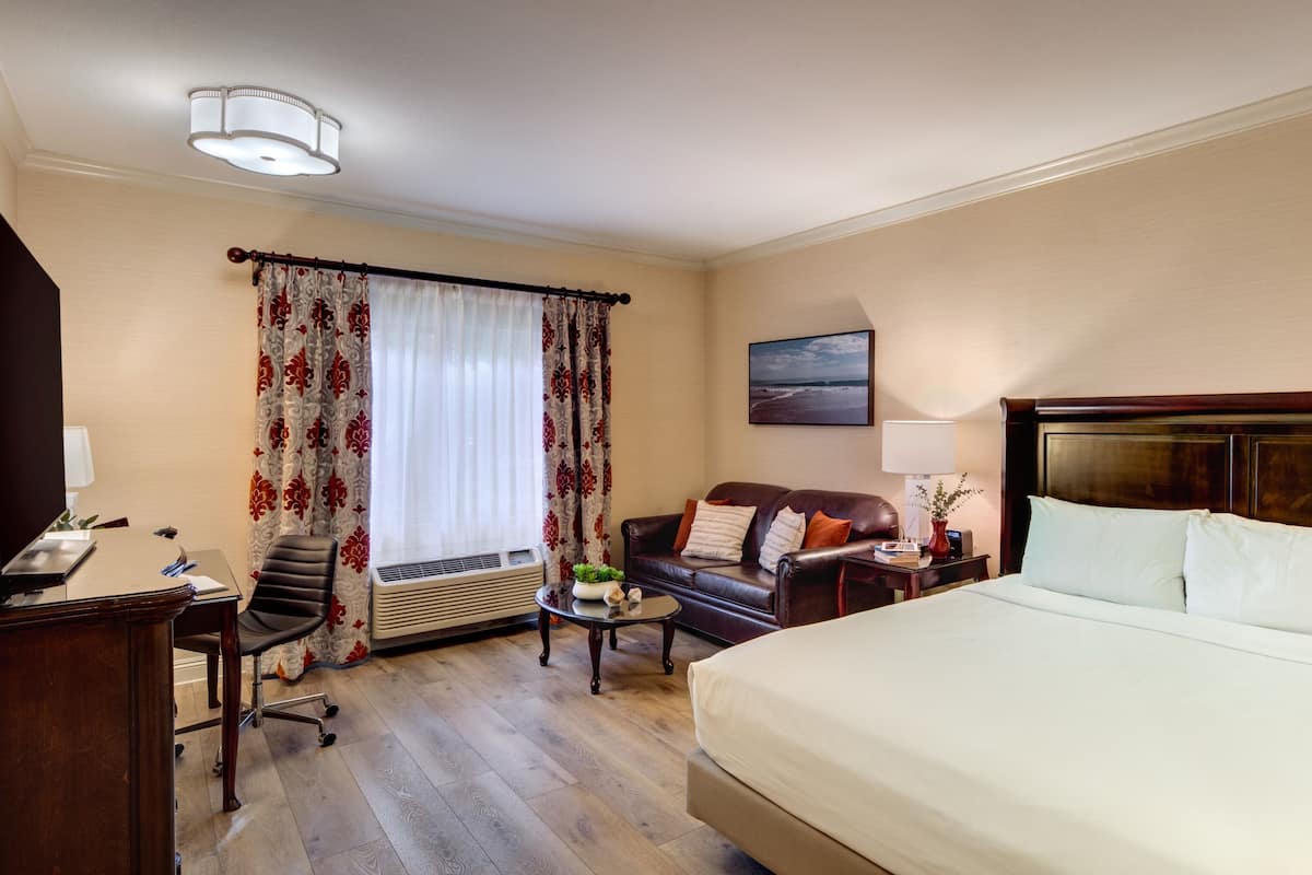 A hotel room with a large bed, a desk with a chair, a sofa, and a window with curtains.