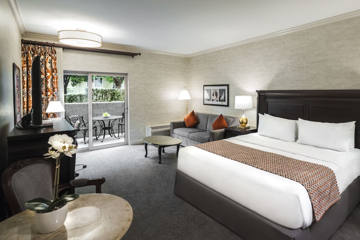 A hotel room with a large bed, a sofa, a dining table, and a chair. There is a window with curtains, a lamp, and a picture on the wall.