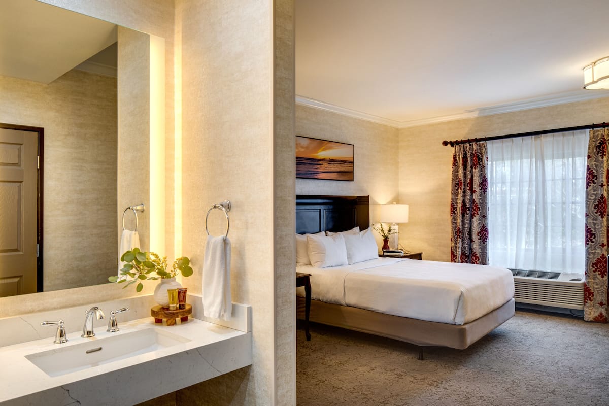 A hotel room with a large bed, a bedside table with a lamp, a window with curtains, and a bathroom with a sink and mirror.