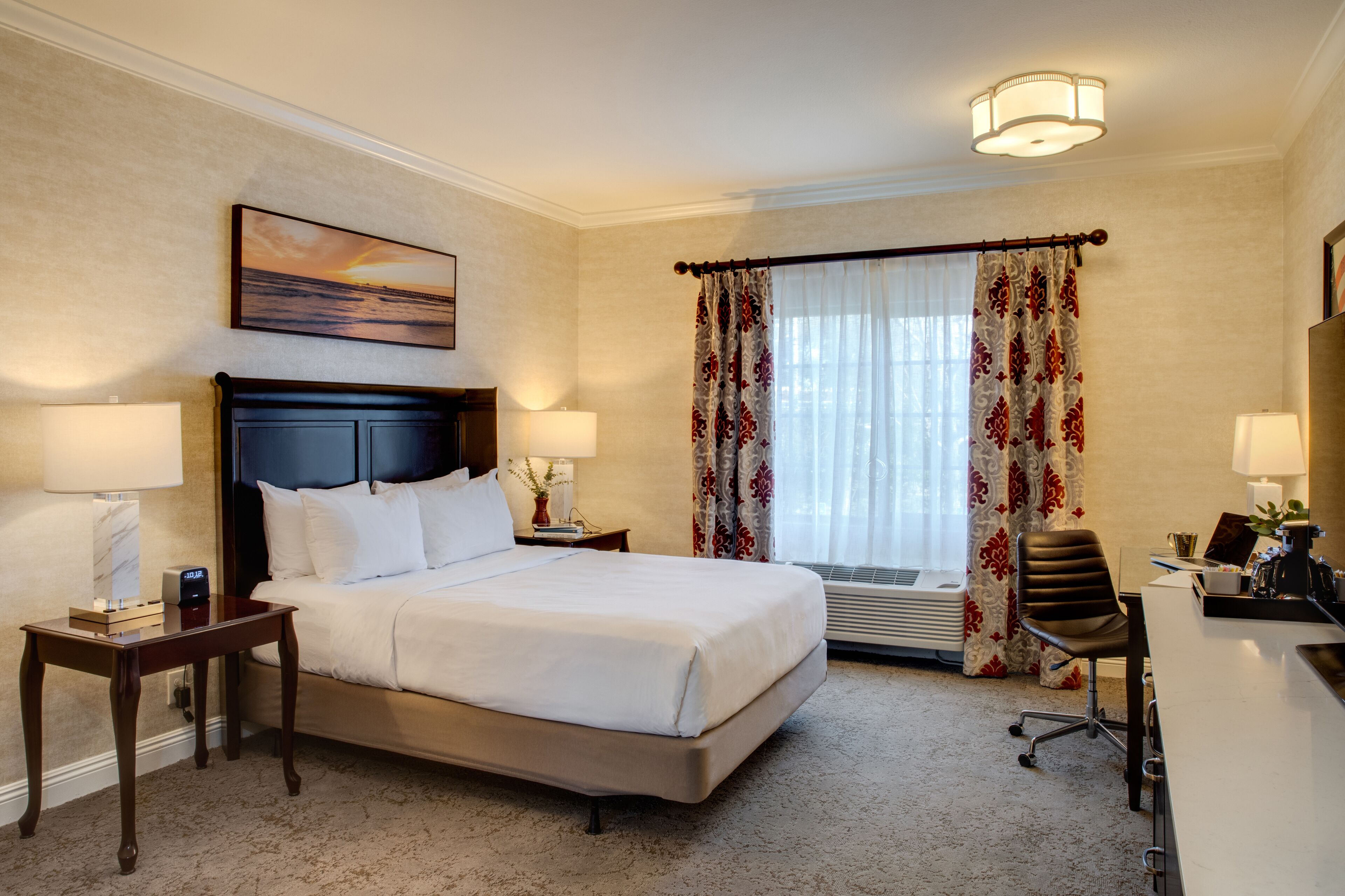 Une chambre d’hôtel avec un grand lit, deux tables de chevet, un bureau avec une chaise, une fenêtre avec des rideaux et un tableau au mur.