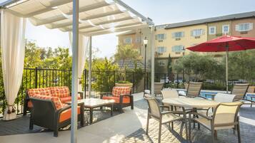 Relax in the shaded patio area, featuring a sofa, chair, and dining tables, surrounded by lush greenery and a pool in the distance.