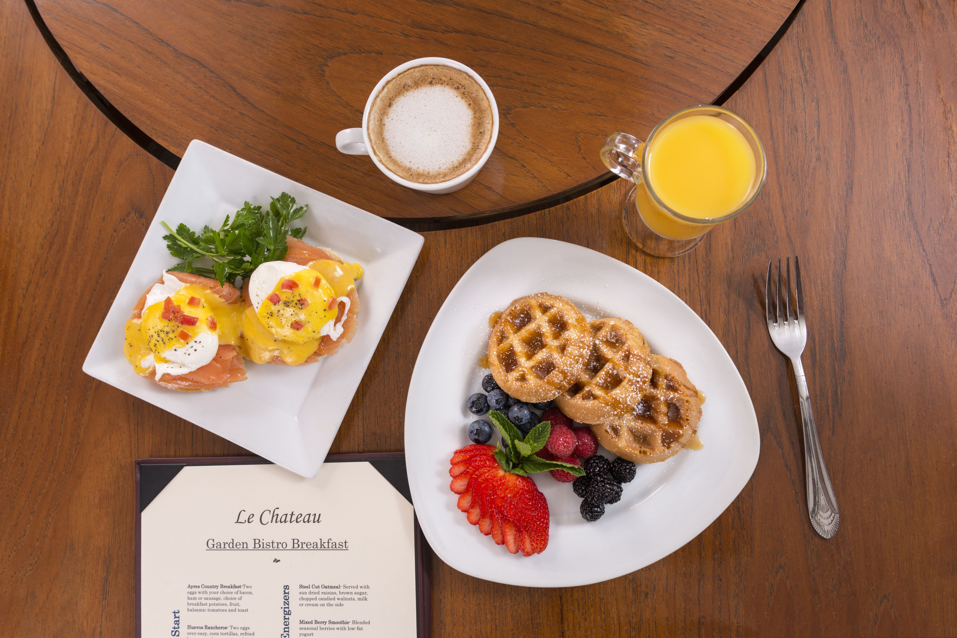 Une table en bois dressée pour le petit-déjeuner, avec un menu, une assiette de œufs Bénédicte, des gaufres et un verre de jus d’orange.