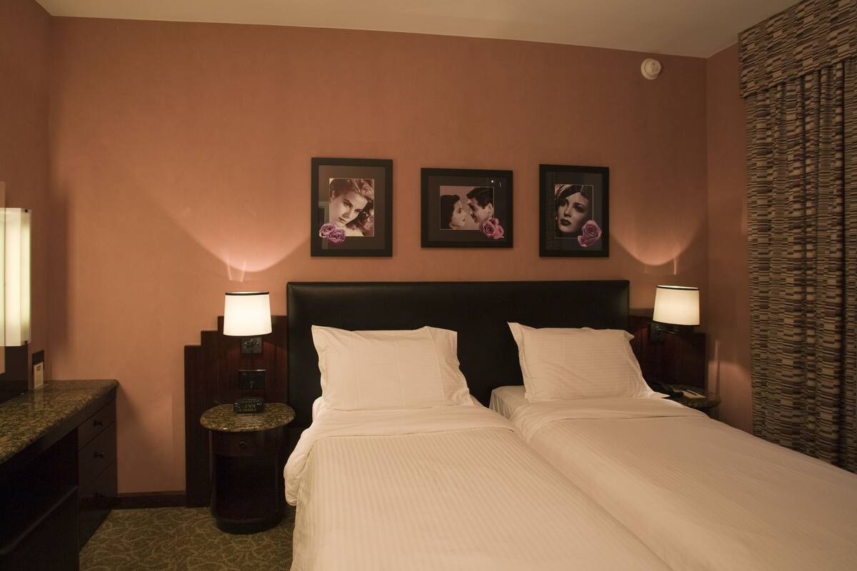 A hotel room with two beds, a nightstand with a clock, a wall clock, and framed pictures on the wall.
