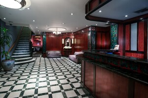 Um hall com piso xadrez, escada, balcão de recepção e um lustre.