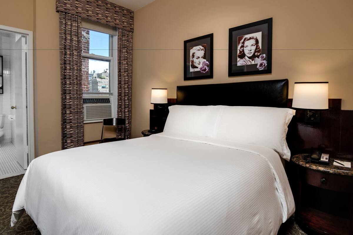 A hotel room with a large bed, two wall-mounted lamps, a window with a view, and two framed pictures on the wall.