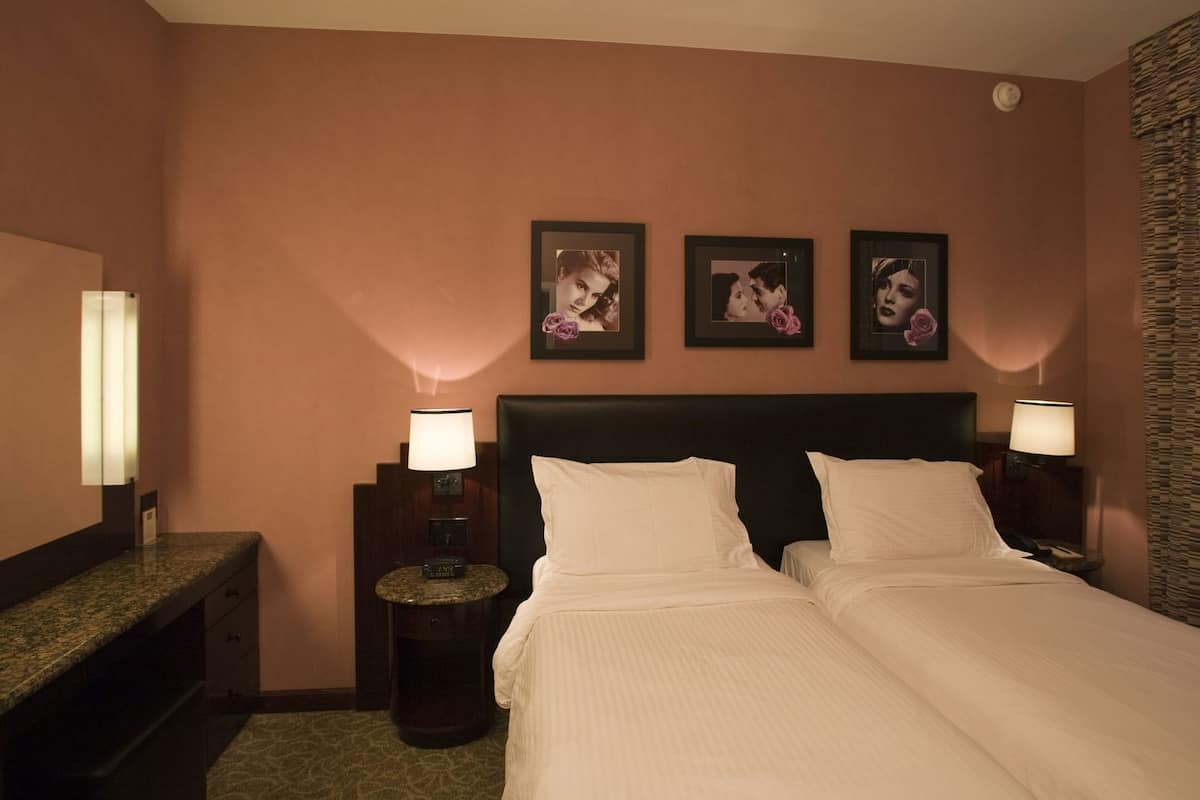 A hotel room with two beds, a headboard, a nightstand with a lamp, a clock, and framed pictures on the wall.