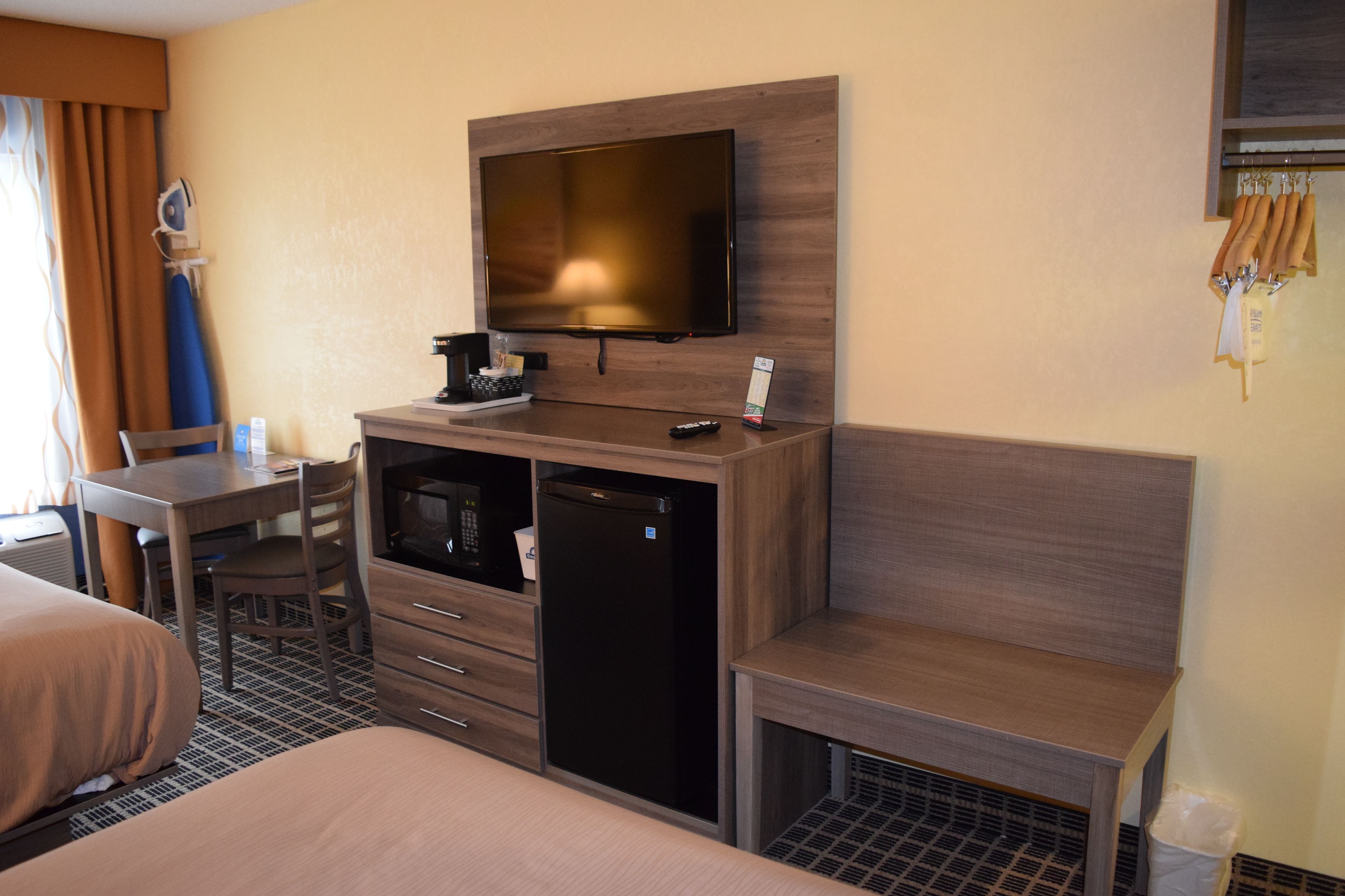 A hotel room with a bed, a desk with a chair, a television mounted on the wall, a mini-fridge, and a trash bin.
