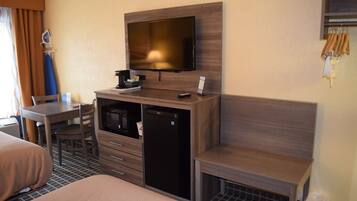 A hotel room with a bed, a desk with a chair, a television mounted on the wall, a mini-fridge, and a trash bin.