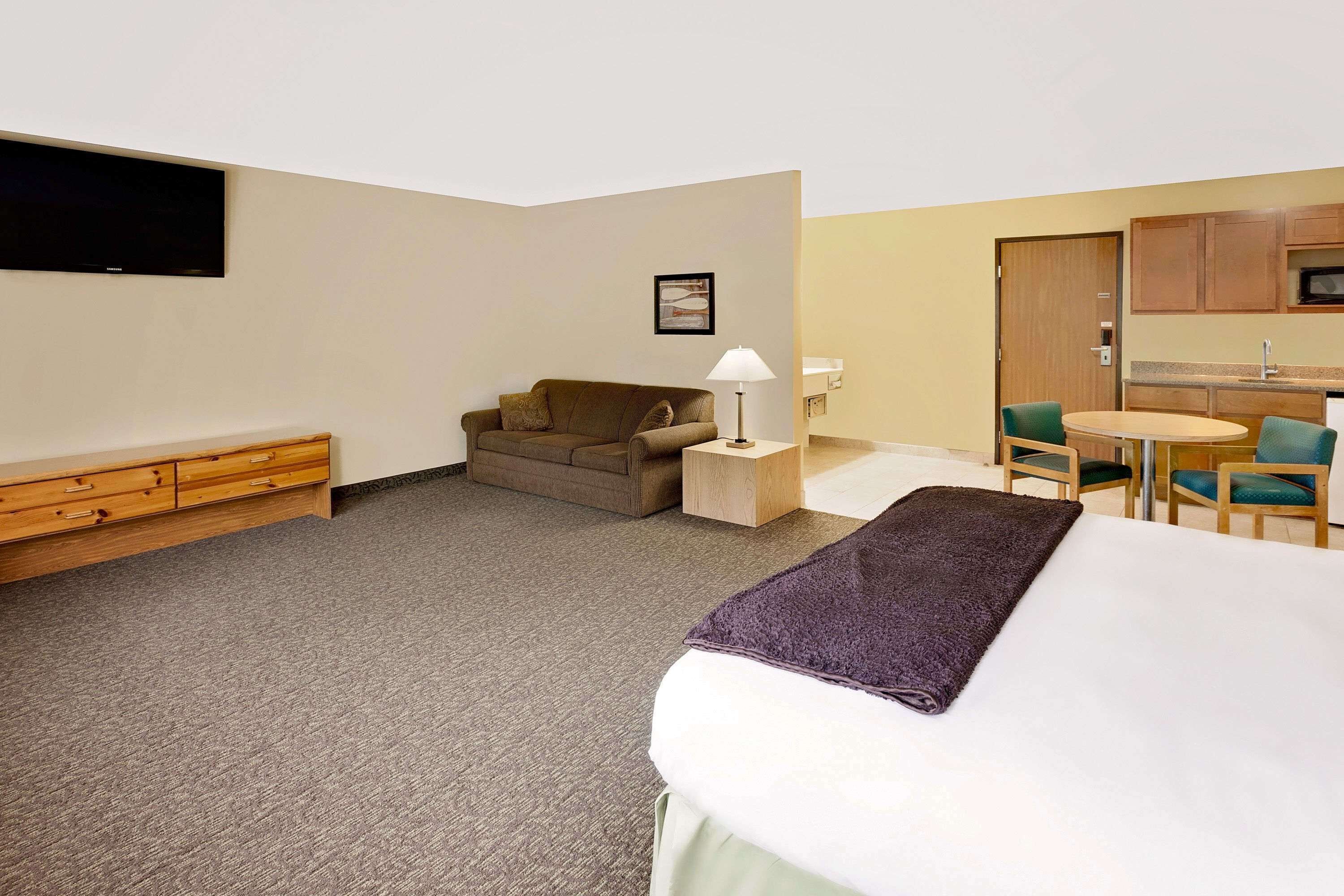 A hotel room with a bed, a sofa, a small table with a lamp, a kitchenette, and a TV mounted on the wall.