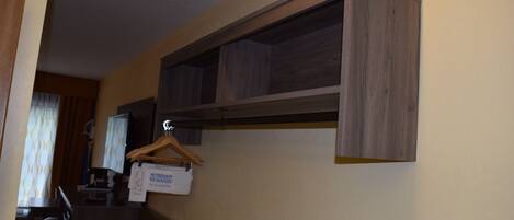 A hotel room with a wooden shelf unit, a desk with a computer, and a hanging garment bag.