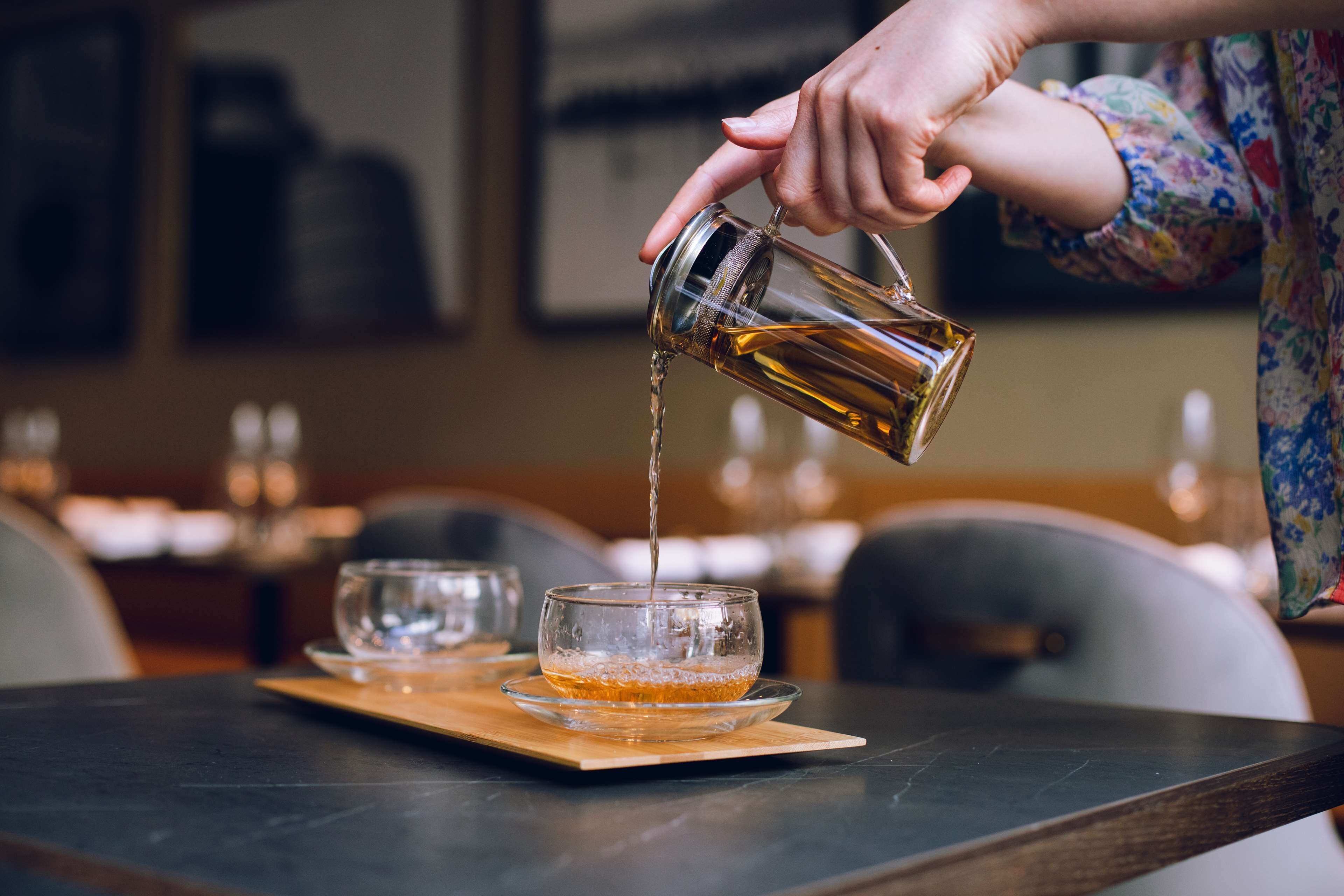 A hand pouring a golden liquid into a clear glass on a wooden tray.