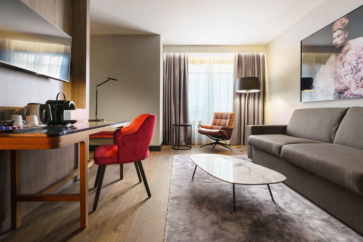 Une chambre d’hôtel moderne équipée d’une télévision à écran plat, d’un bureau avec une machine à café, d’un fauteuil rouge, d’un canapé gris et d’un grand tableau accroché au mur.