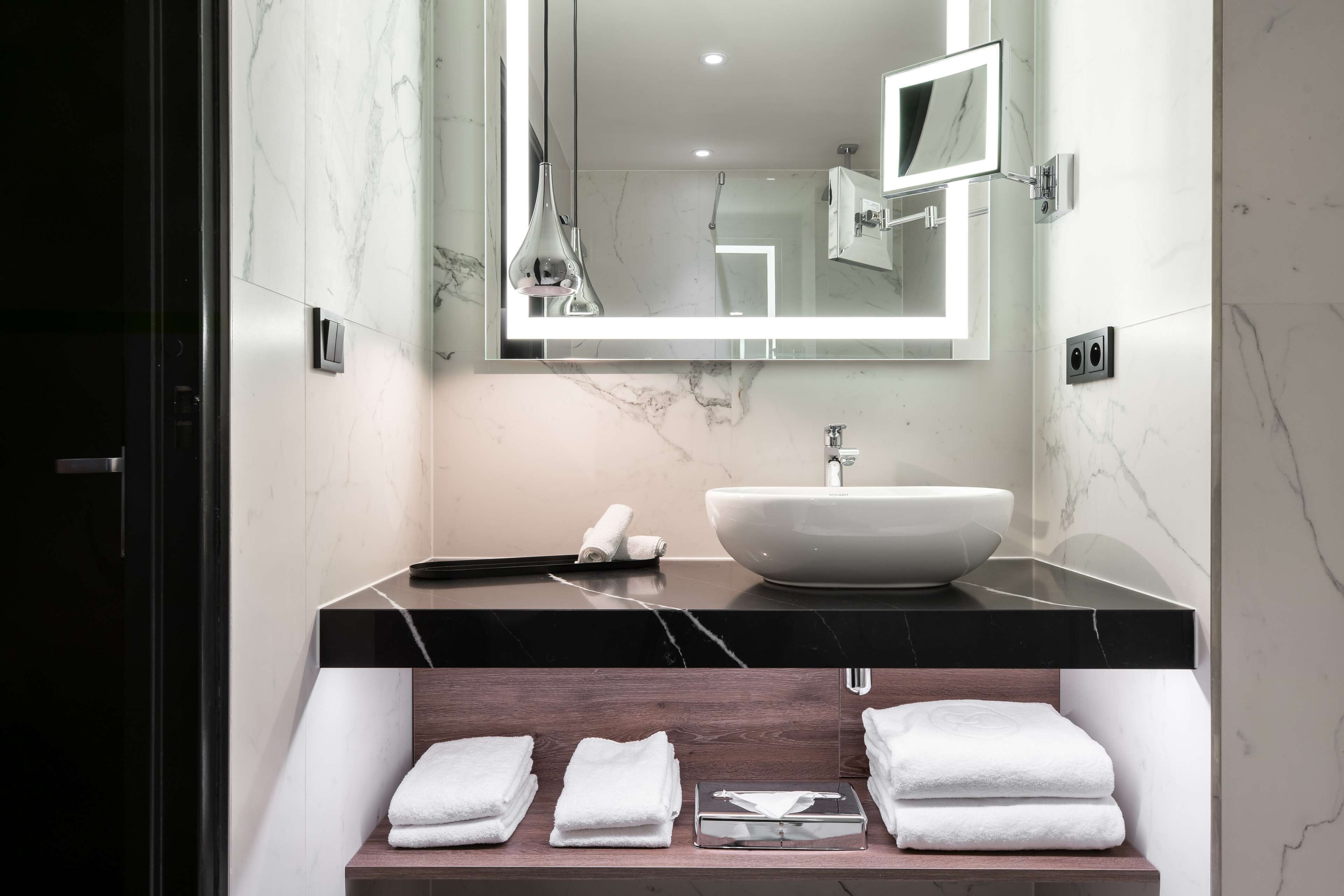 A modern bathroom with a large mirror, a white vessel sink, and neatly folded towels on a wooden shelf.