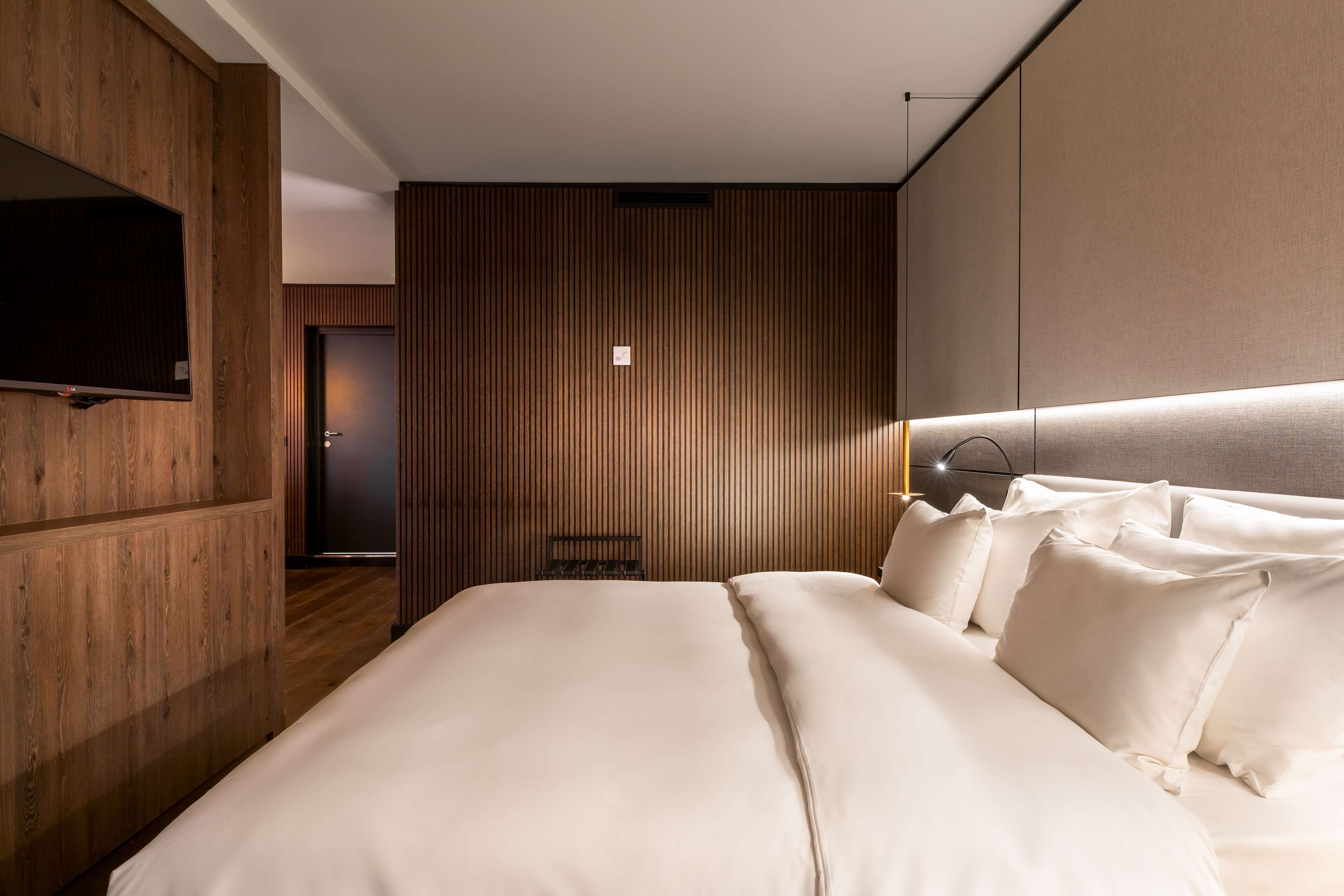 A modern hotel room with a large bed, wooden paneling, a flat-screen TV, and a bedside table.