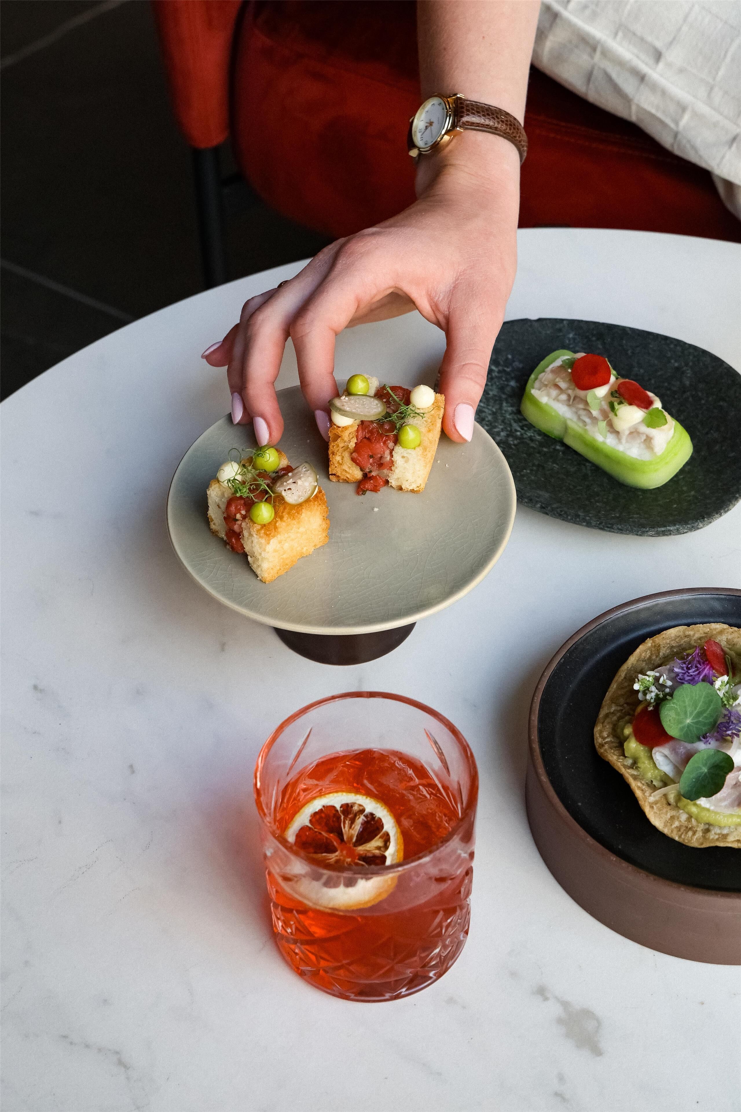 A person serving food on a round table with a plate of appetizers, a glass of red drink, and a small bowl of salad.