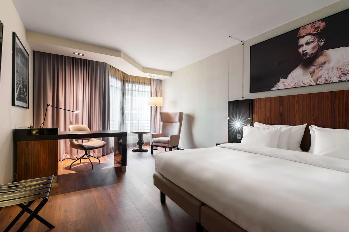 Une chambre d’hôtel avec un grand lit, un bureau avec une chaise, une chaise supplémentaire, une lampe et un grand tableau encadré au mur.