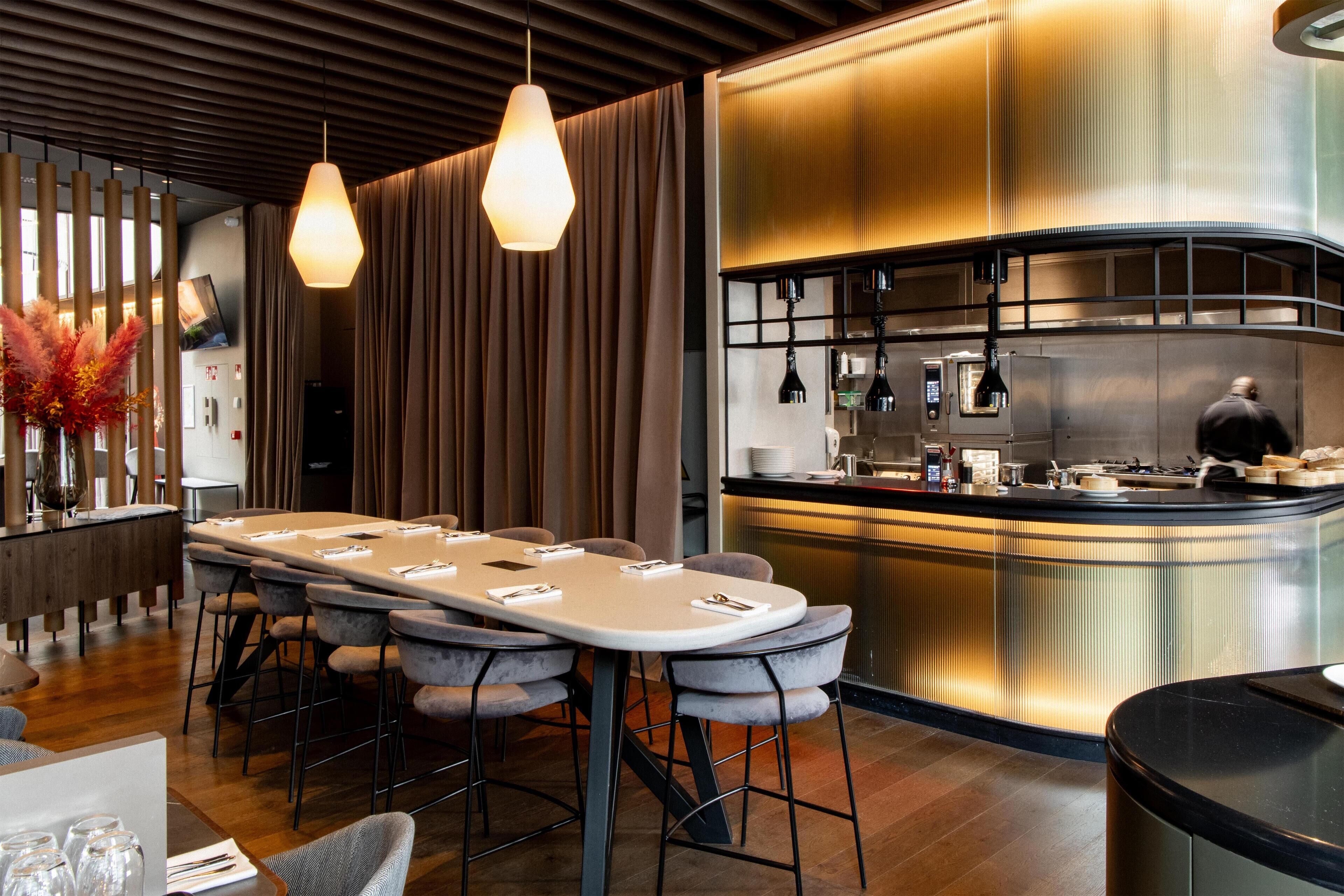 A modern restaurant with a long communal table, bar stools, a bar area, and a kitchen in the background.