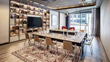 A conference room with a long table, chairs, a large flat-screen TV, and a bookshelf filled with books and decorative items.