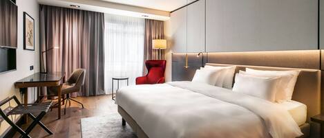 A hotel room with a large bed, a desk, a red chair, and a lamp.
