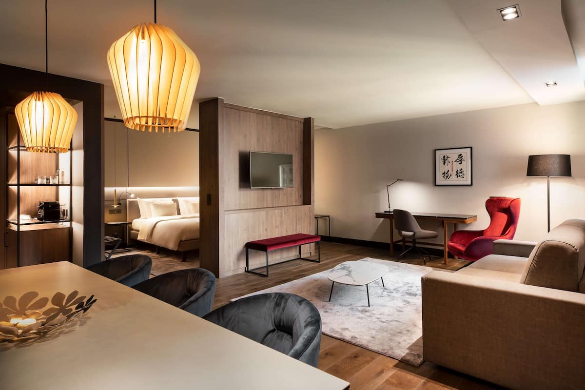 Une chambre d’hôtel moderne avec un plancher en bois, un téléviseur à écran plat fixé au mur, un banc rouge, un bureau avec une chaise et un canapé.
