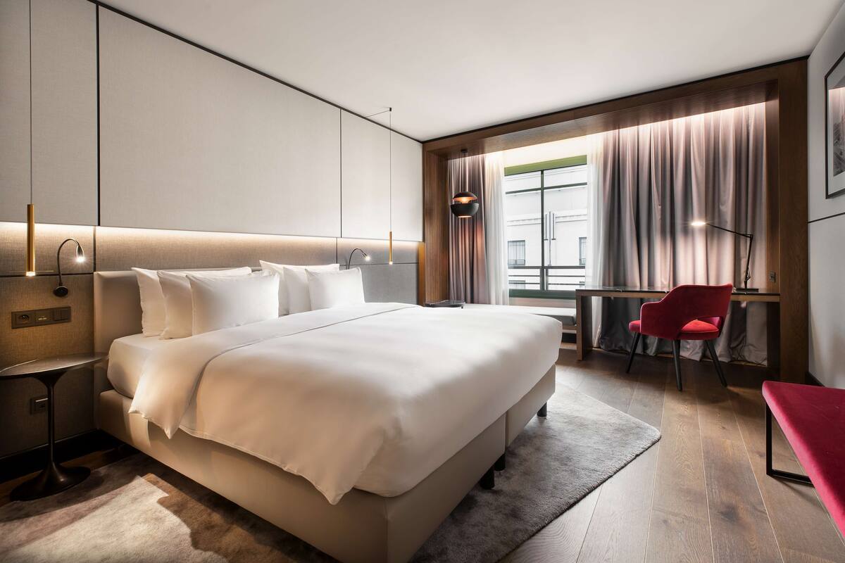 Une chambre d’hôtel moderne dotée d’un grand lit, d’un bureau, d’une chaise et offrant une vue sur la ville grâce à de grandes fenêtres.