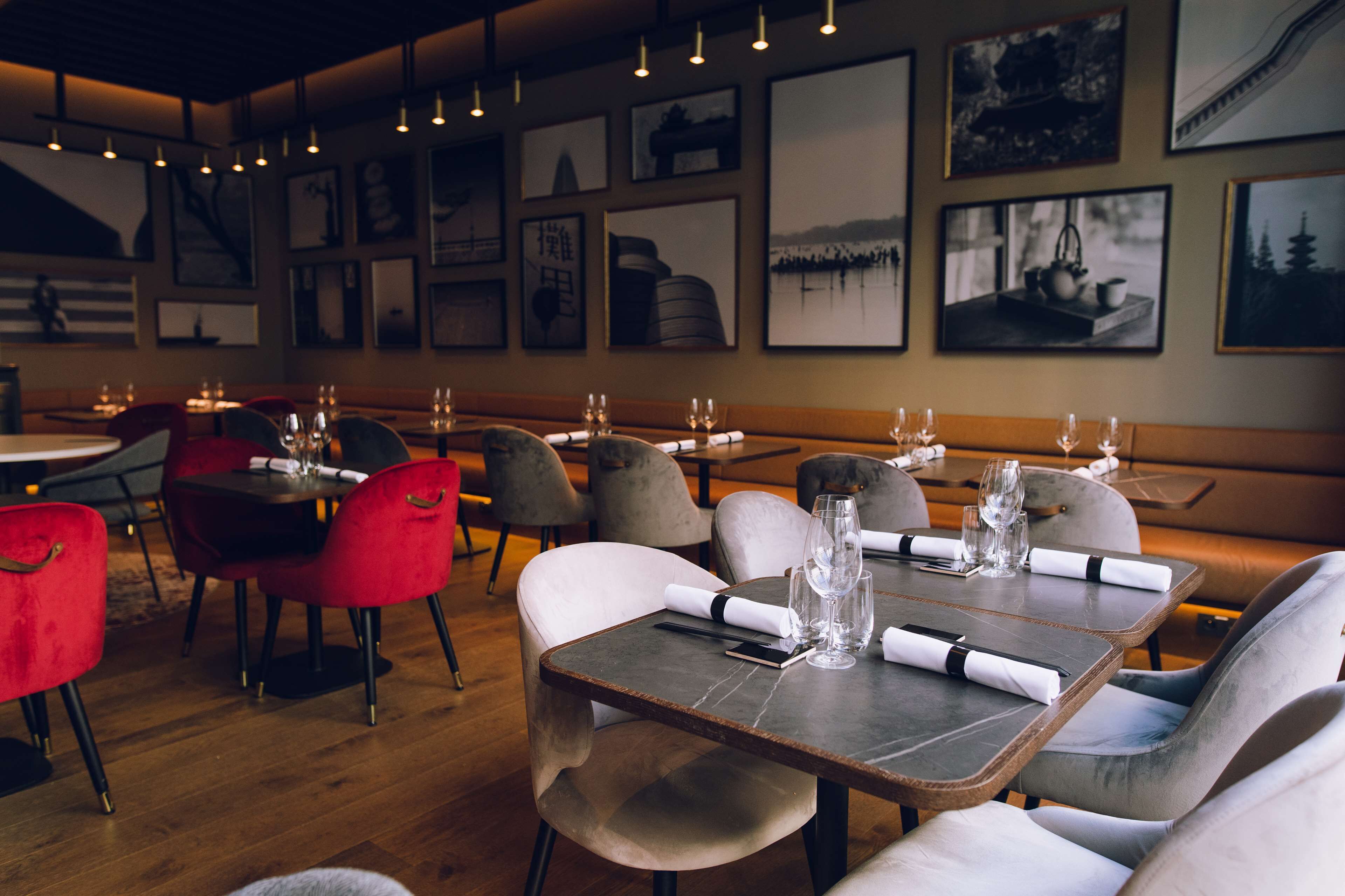 Interior de restaurant amb taules de fusta, cadires tapissades de vermell i gris, i quadres a les parets.