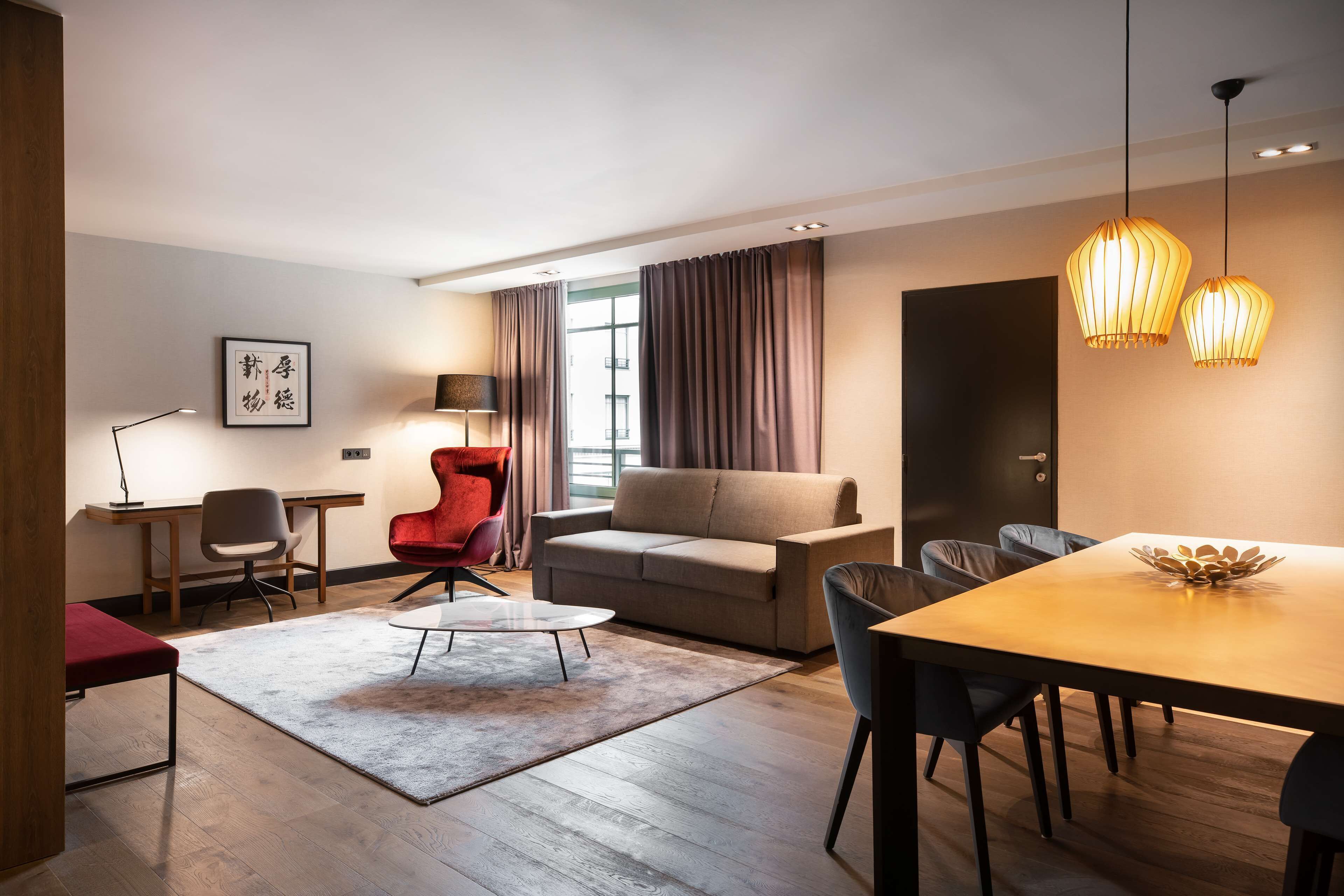 Una sala moderna con sofá, sillón rojo, mesa de comedor y lámparas colgantes.