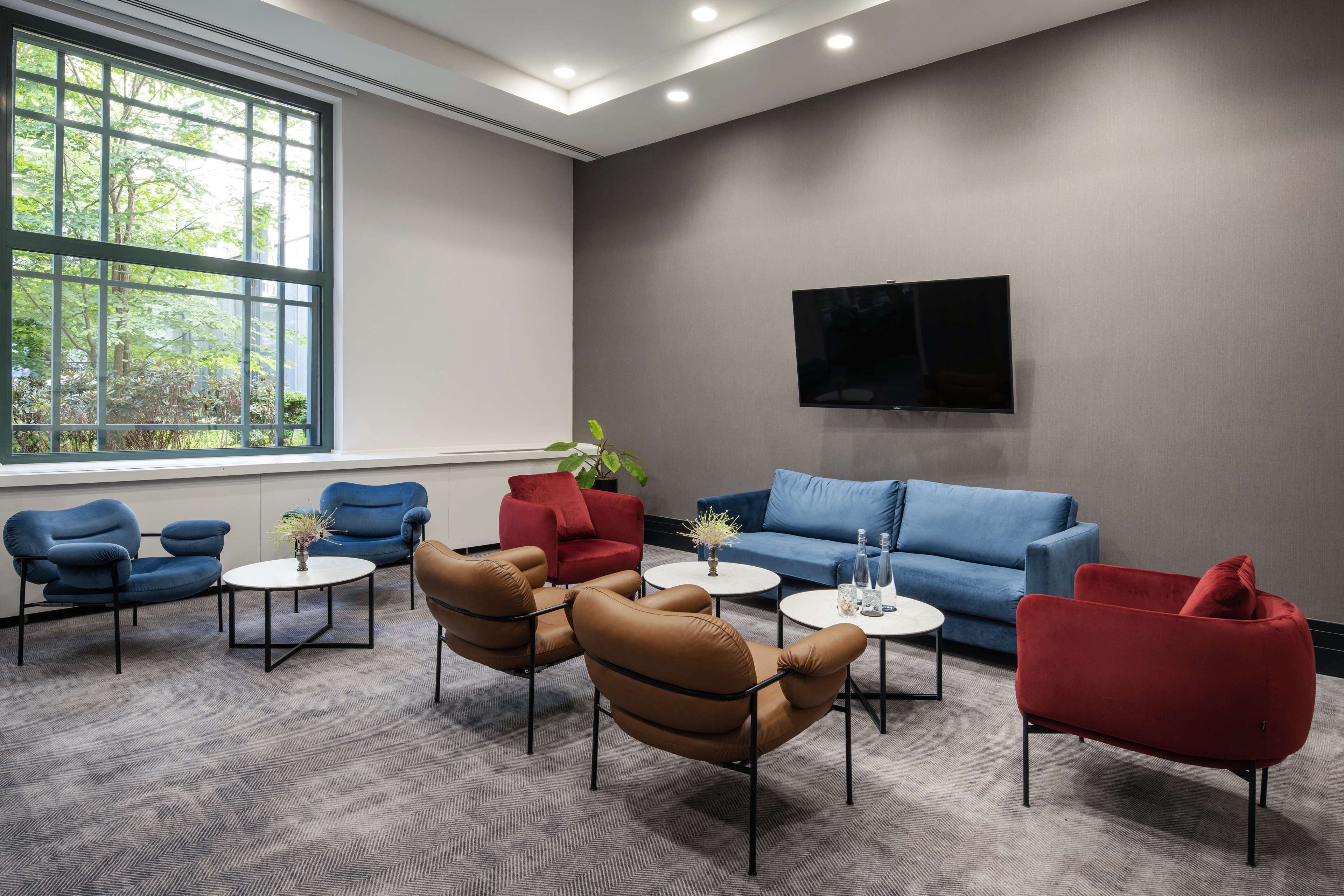 A modern lounge area with a grey carpet, blue and red armchairs, a blue sofa, and a flat-screen TV on the wall.