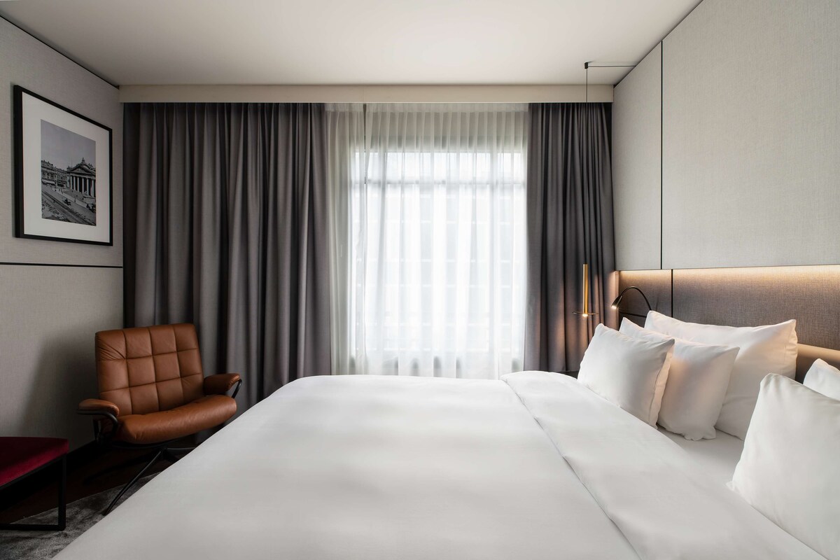 Une chambre d’hôtel avec un grand lit, une chaise, un tableau encadré au mur et une fenêtre avec des rideaux.